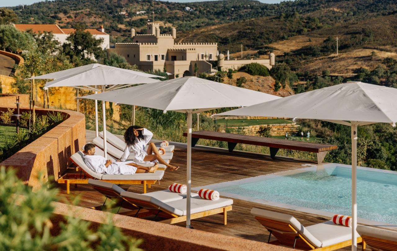 Terraço da piscina do resort com espreguiçadeiras e guarda-sóis, tendo como pano de fundo a arquitetura histórica.
