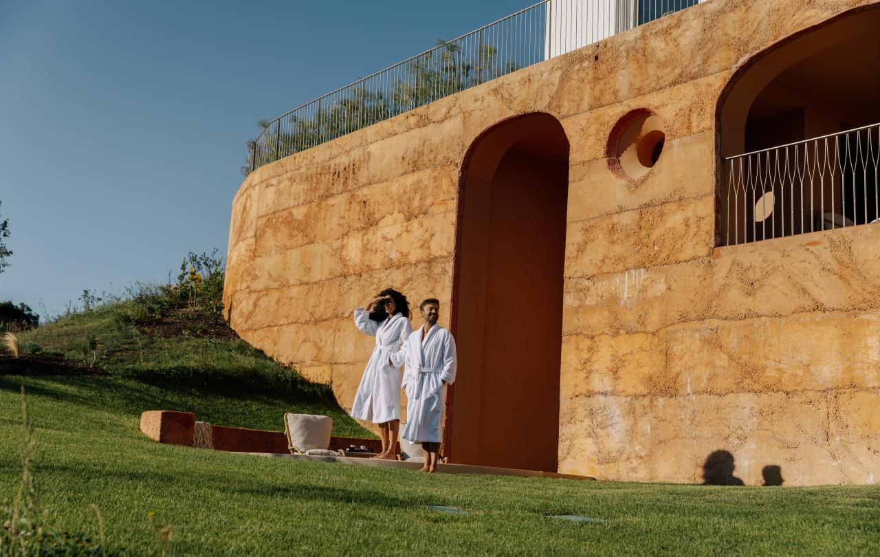 Casal com roupões brancos, em pé junto a um edifício singular de terracota, num relvado verdejante.