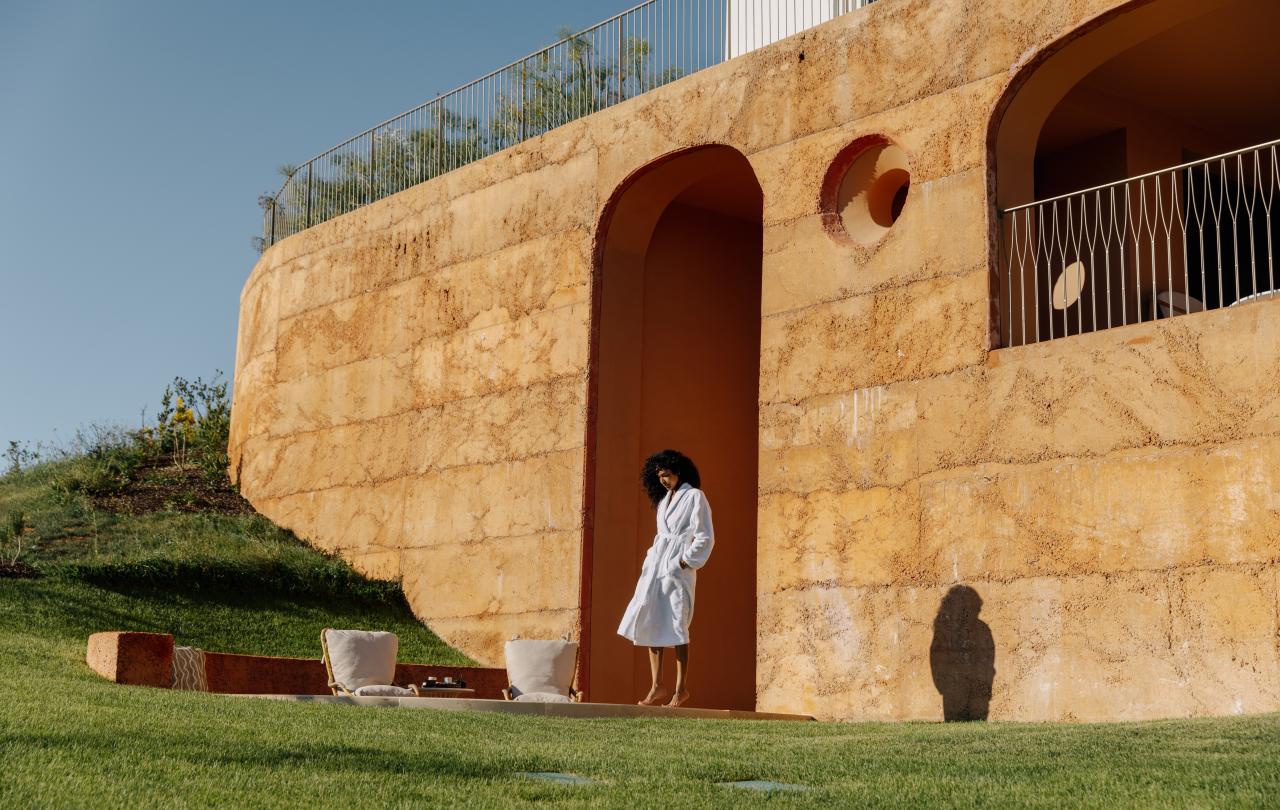 Mulher vestida com um roupão branco, em pé junto a um edifício singular de terracota, num relvado verdejante.