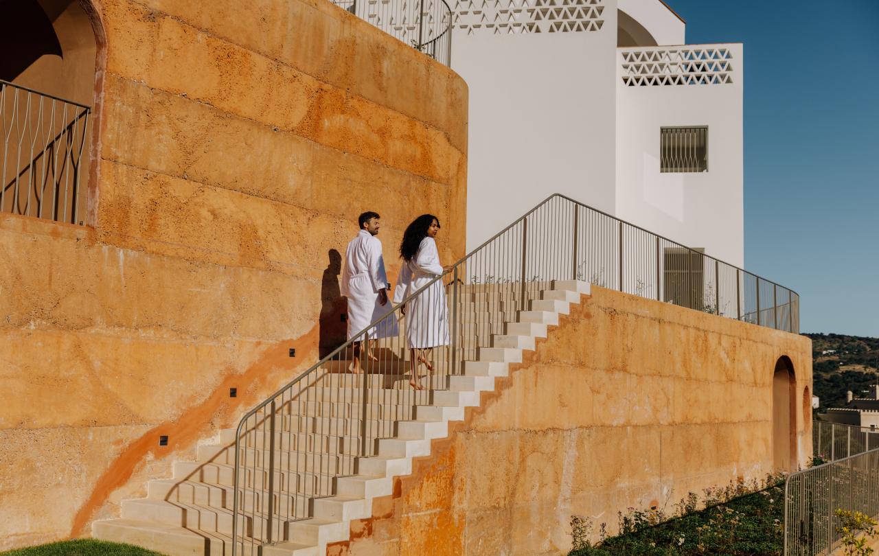 Casal com roupões brancos a subir uma escadaria exterior de pedra ao lado de um edifício em tons quentes.