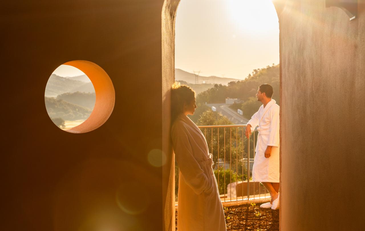 Casal de roupão, debaixo da luz quente do sol, junto a um arco com vista para o campo.