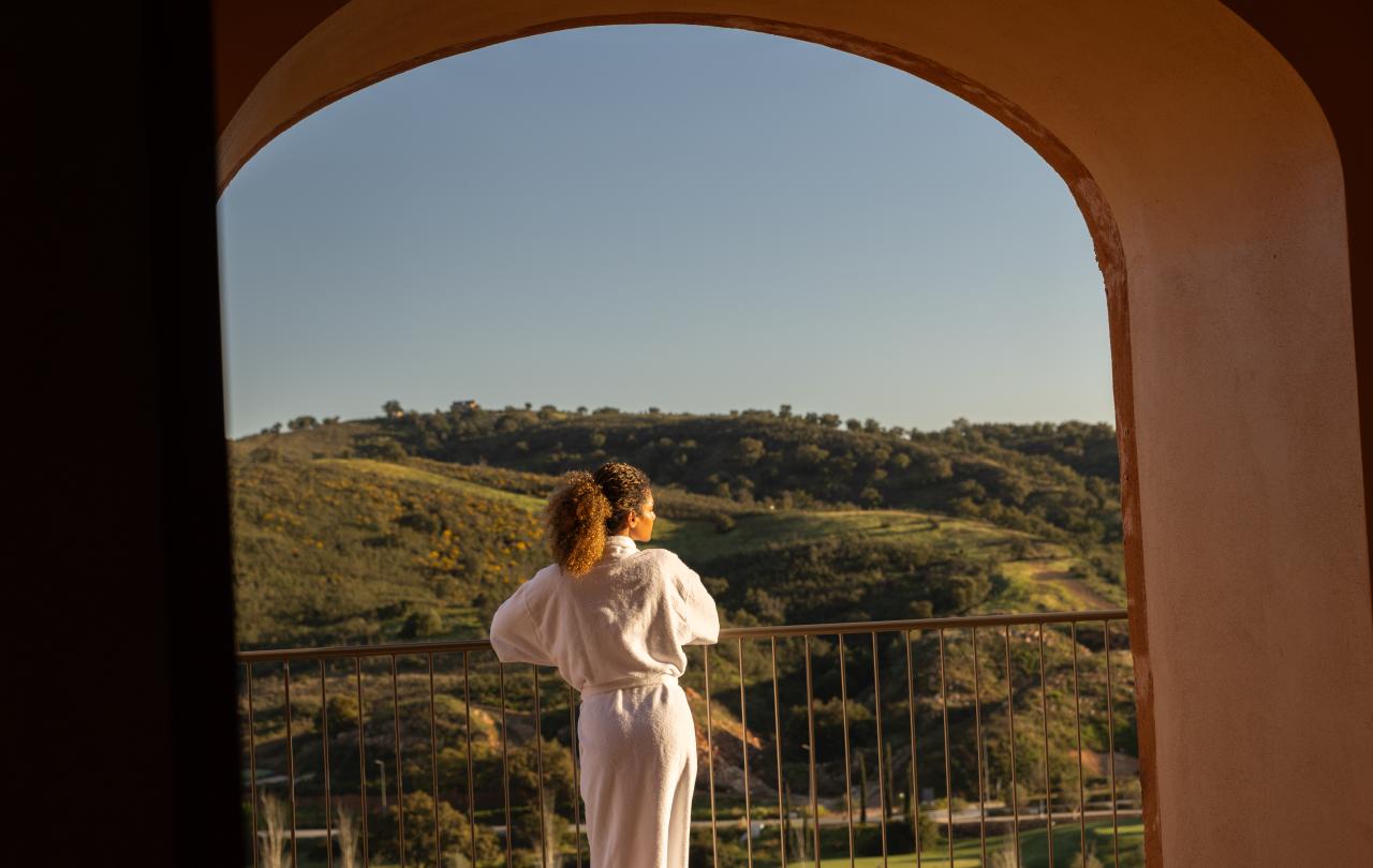 Mulher vestida com um roupão branco a contemplar as colinas verdes através da janela arqueada da varanda.
