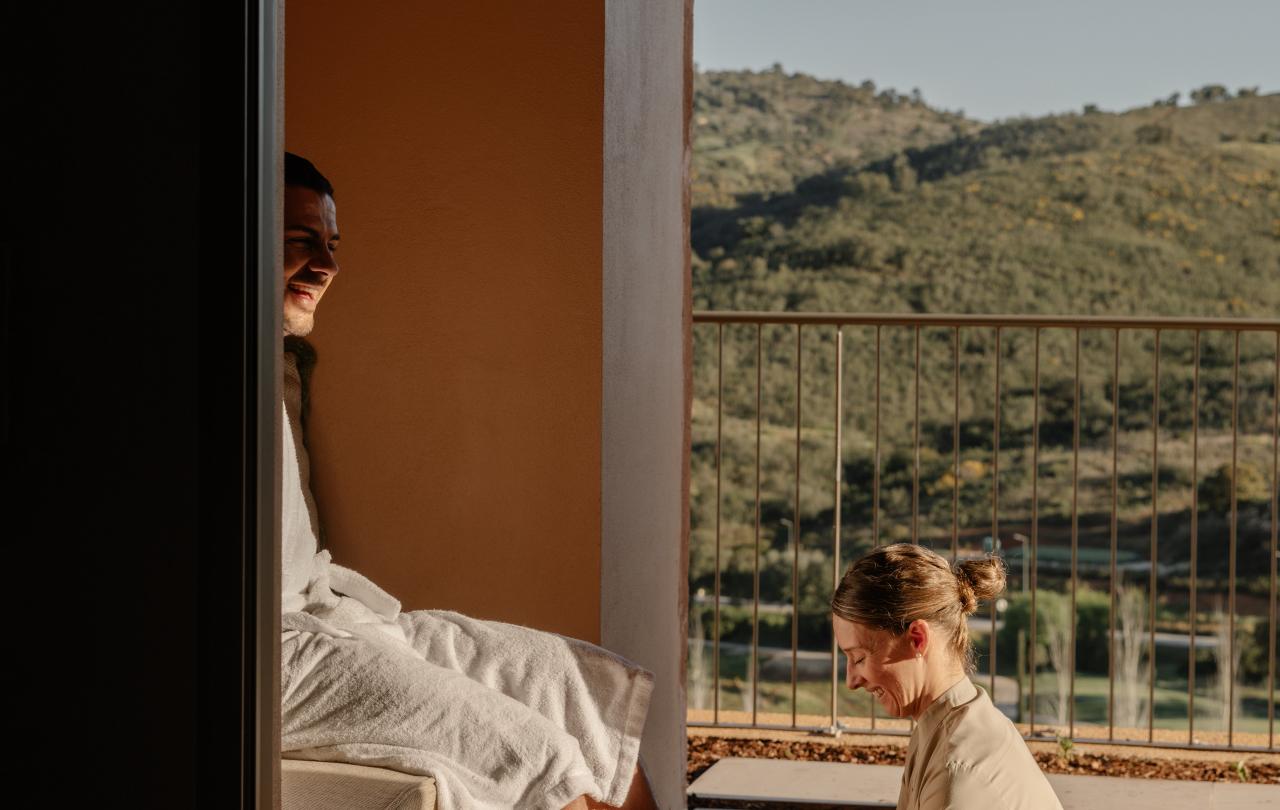 Um profissional de spa a realizar um tratamento relaxante de banho de pés a um hóspede num terraço ensolarado.