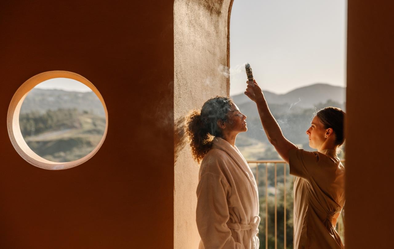 Um terapeuta de spa a purificar um hóspede, vestido com um roupão de banho, com um pau de sálvia aceso, num terraço.