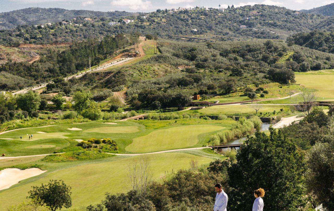 Casal com roupões brancos, junto à beira de uma piscina infinita com vista para um extenso campo de golfe.