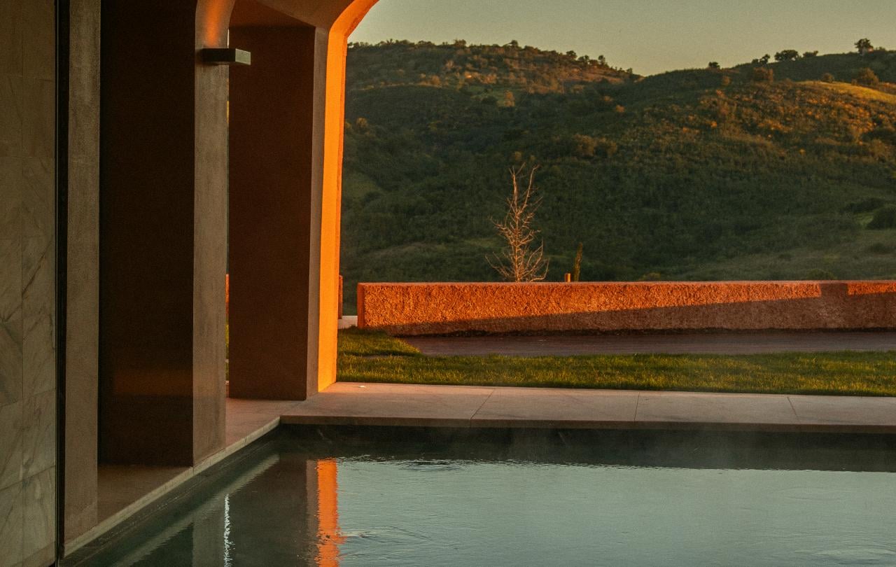 Piscina de spa interior sob um arco de pedra, refletindo a luz quente do pôr-do-sol e as colinas ondulantes lá fora.