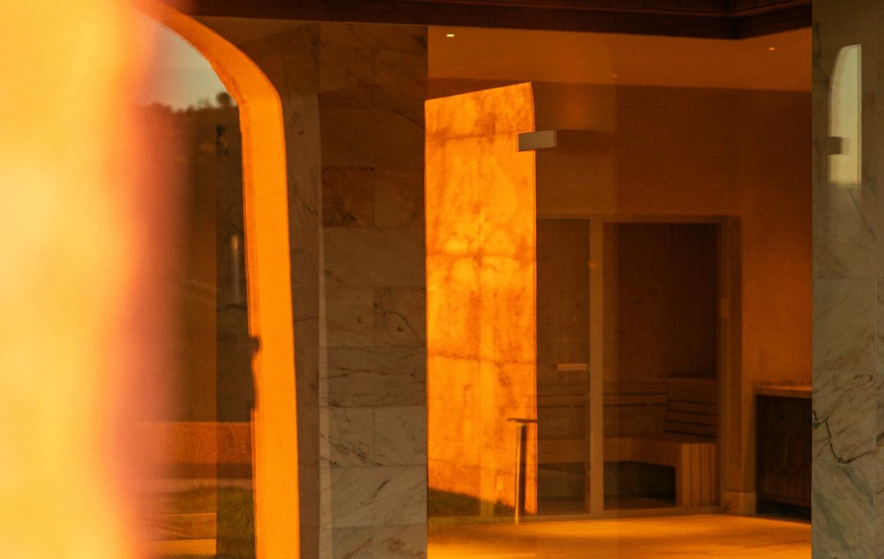 Um brilho dourado e acolhedor ilumina uma luxuosa área de piscina interior e a sauna, que se refletem na água.