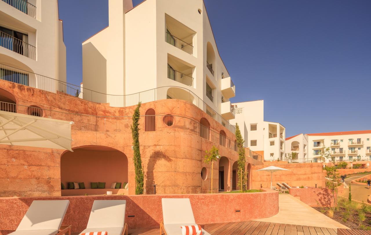 Os edifícios brancos com varandas têm vista para um terraço em tons de terracota, com espreguiçadeiras e um deck de madeira.