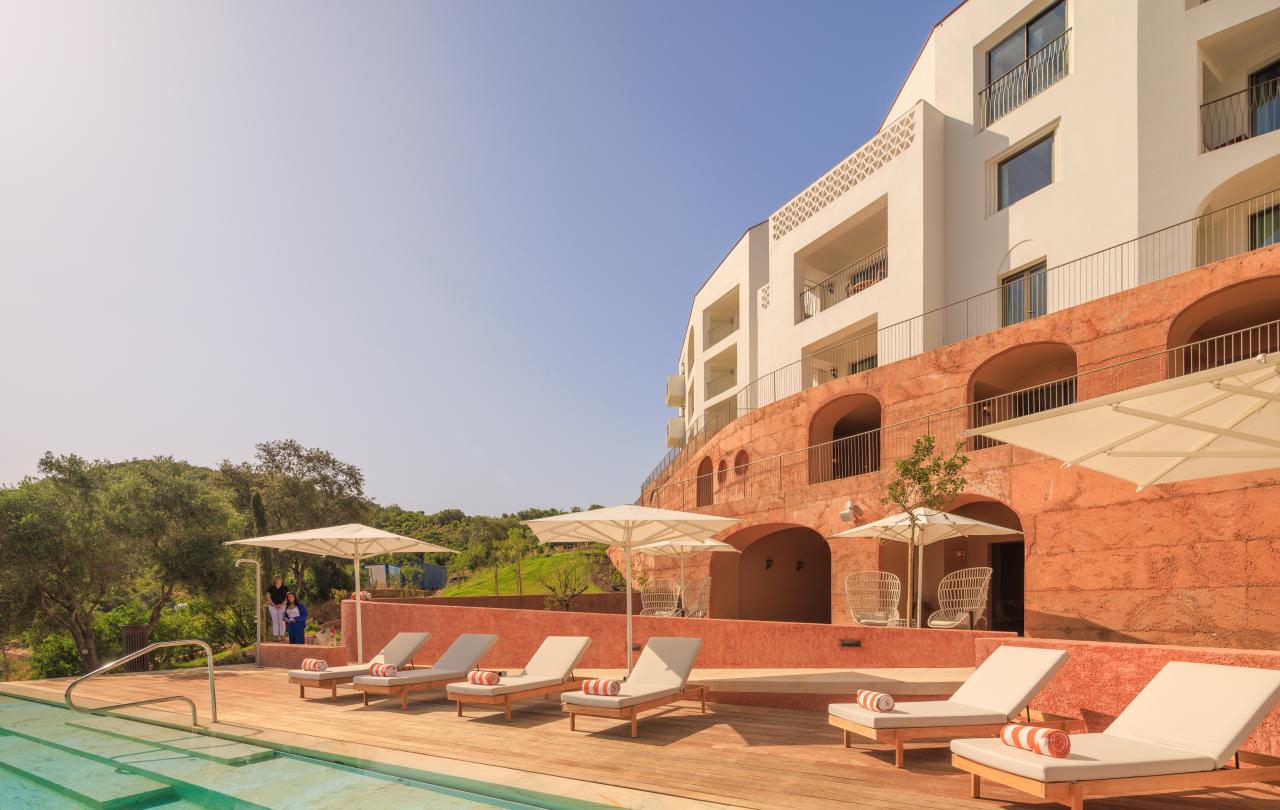 Um moderno edifício de hotel branco, com entradas em arco e varandas, tem vista para uma piscina e espreguiçadeiras num dia de sol.