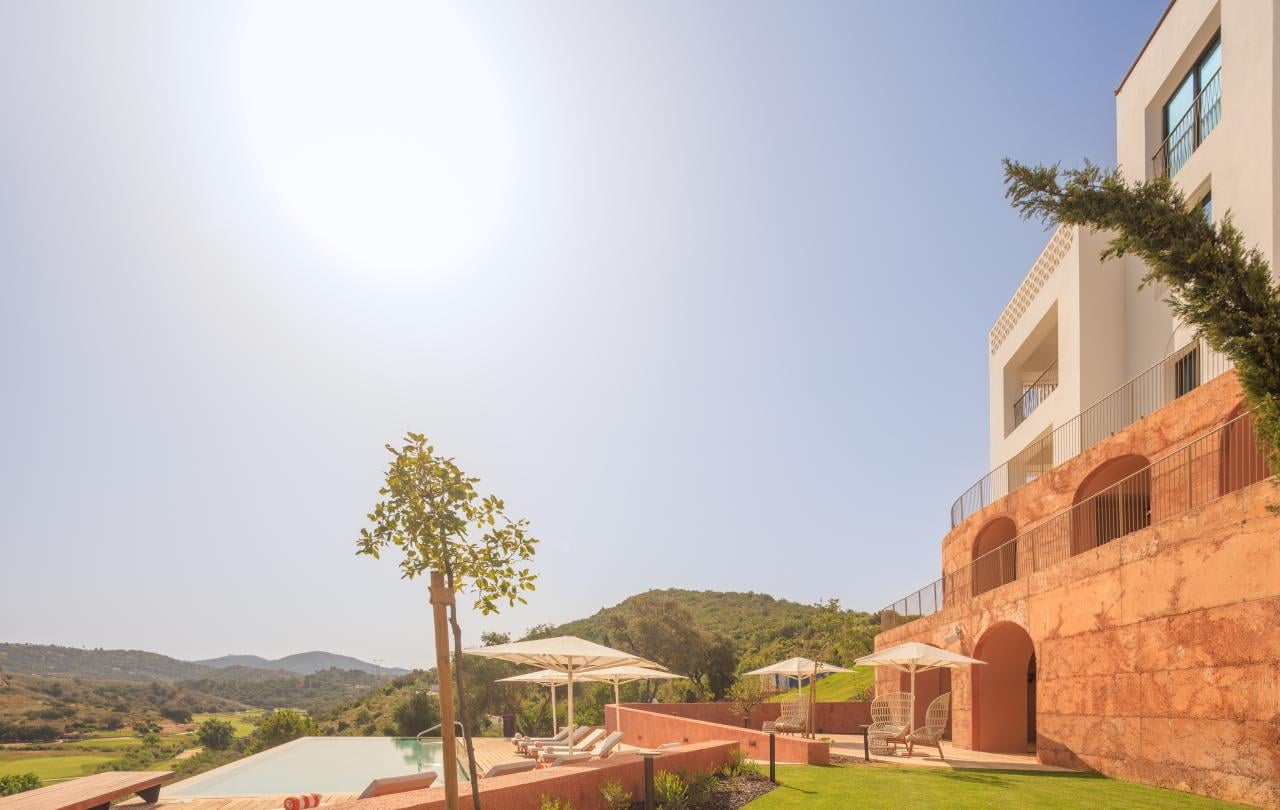 Edifício hoteleiro moderno e branco, com uma base em terracota com arcos, com vista para uma paisagem pitoresca, com uma piscina infinita e espreguiçadeiras.