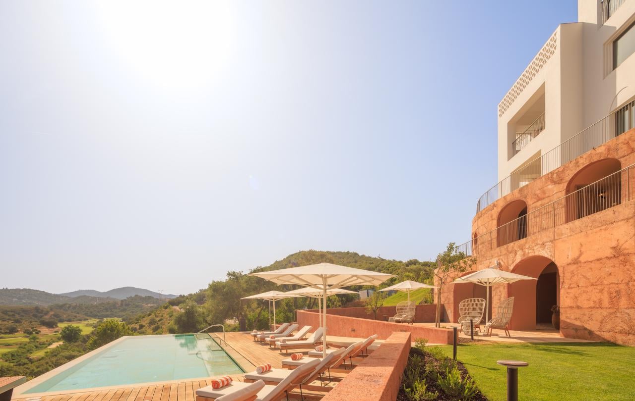 Resort de luxo com piscina infinita, espreguiçadeiras e uma arquitetura hoteleira moderna, inserido numa paisagem montanhosa.