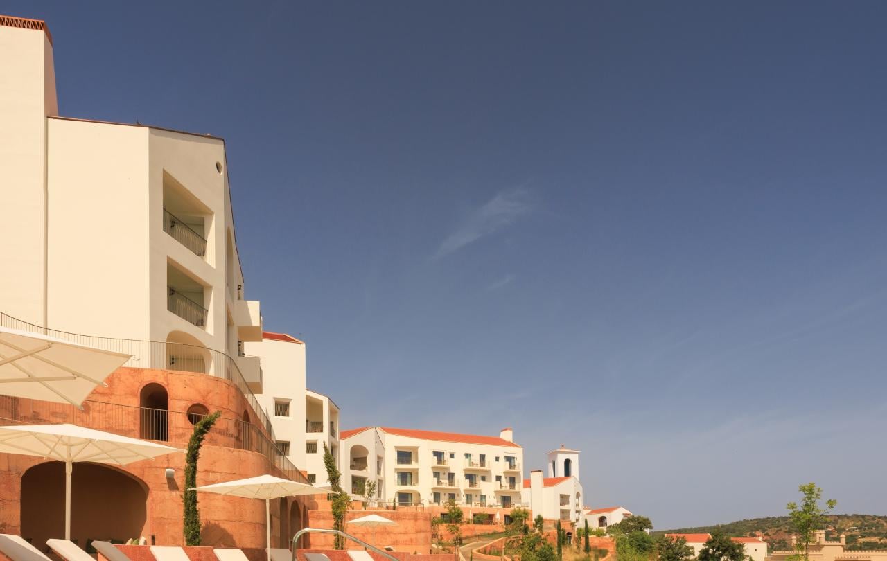 Resort de luxo com uma piscina infinita, espreguiçadeiras brancas e edifícios brancos de vários andares sob um céu azul límpido.