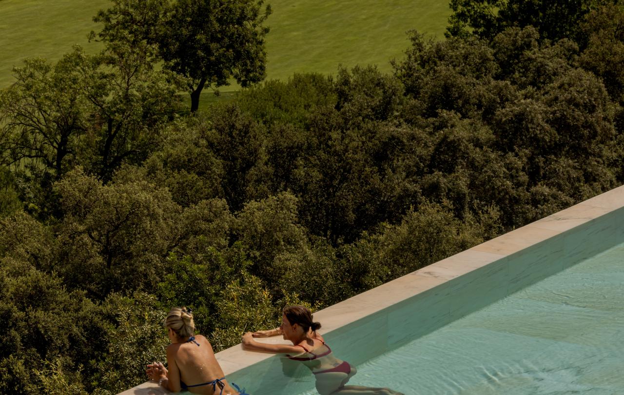 Duas mulheres relaxam numa piscina infinita com vista para um campo de golfe verdejante, onde se avistam carrinhos de golfe e jogadores de golfe ao longe.