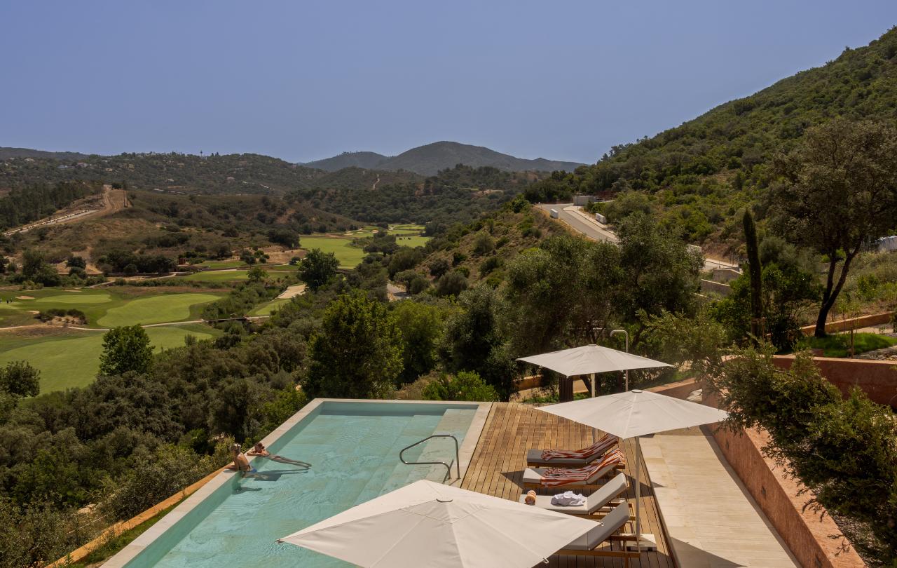 A piscina infinita, com espreguiçadeiras e guarda-sóis, tem vista para um campo de golfe e colinas ondulantes, sob um céu azul límpido.