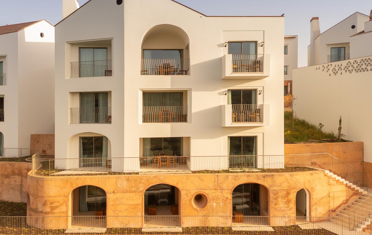 Edifícios modernos e brancos de um resort, com varandas em arco e pisos inferiores em pedra dispostos em degraus, que incluem uma piscina e espreguiçadeiras.