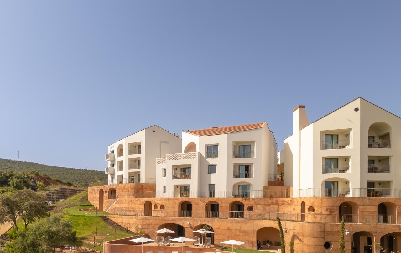 Edifícios modernos e brancos, com terraços em arco e uma piscina, estão aninhados numa encosta, sob um céu azul límpido.