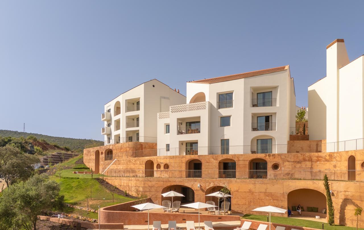 Edifícios modernos brancos, com janelas em arco e varandas, têm vista para uma paisagem em socalcos com uma piscina e espreguiçadeiras.