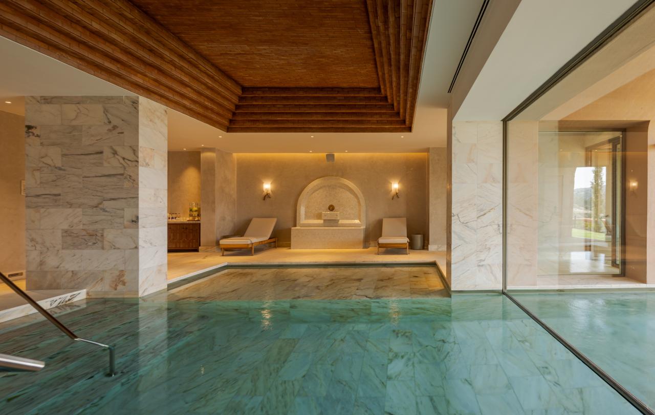 Piscina interior luxuosa com águas cristalinas de cor turquesa e paredes de mármore, com espreguiçadeiras sob um teto de madeira em degraus.