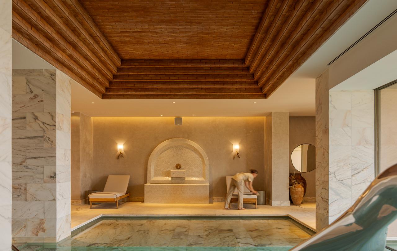 Piscina coberta com azulejos turquesa, situada num spa moderno e luxuoso com colunas de mármore, um teto em degraus de madeira e espreguiçadeiras ao fundo.