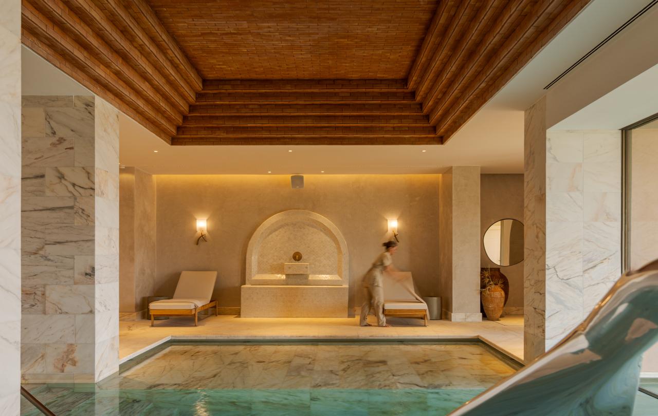 Um spa interior de luxo com uma piscina de mármore turquesa, espreguiçadeiras e um teto de madeira em camadas, visto através de uma grade metálica em primeiro plano.