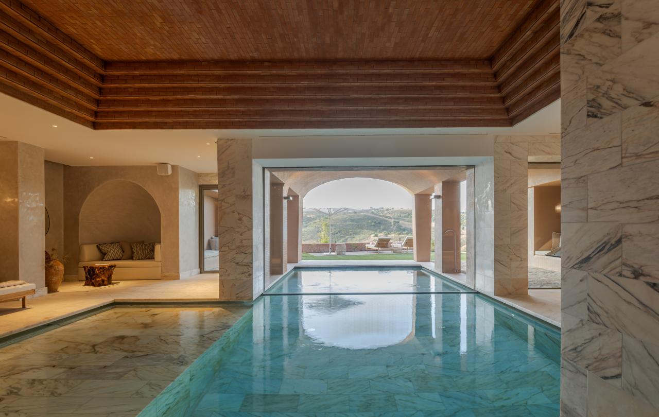 Uma piscina interior com azulejos de mármore e um teto em caixotões de madeira oferece uma vista para uma paisagem ondulante através de grandes janelas.