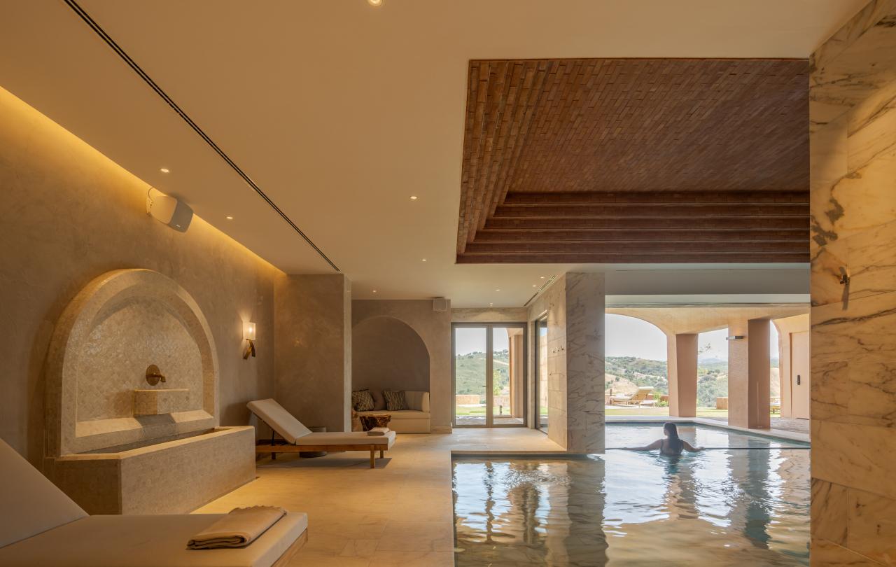 Spa interior de luxo com uma fonte de mármore, espreguiçadeiras de madeira e uma piscina interior-exterior, com uma pessoa a nadar ao fundo.