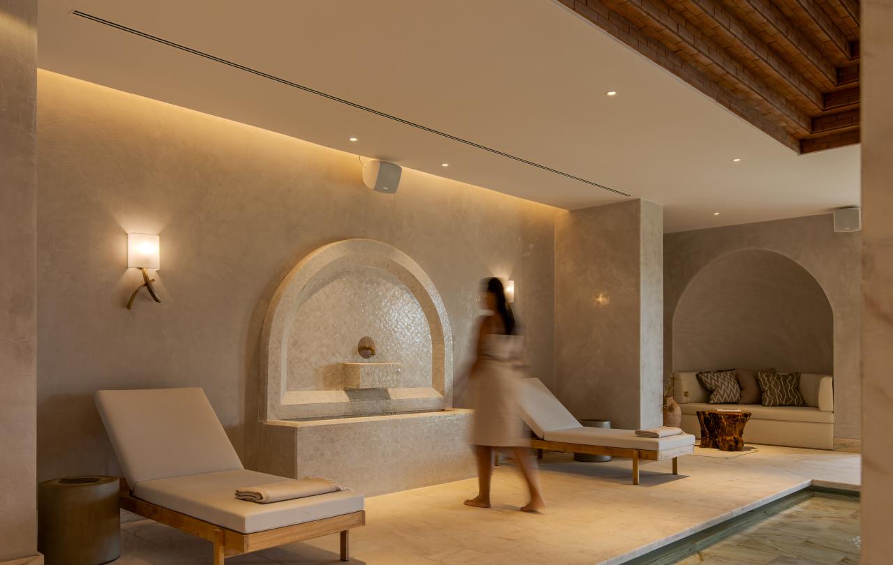 Uma pessoa vestida com um roupão passa por uma fonte de pedra e por pessoas a relaxar numa sala de spa bem iluminada, com uma piscina revestida a azulejos.