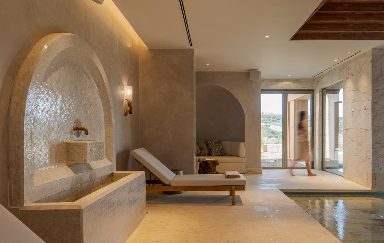 Área moderna de piscina interior com espreguiçadeiras, paredes de pedra, uma fonte decorativa e grandes janelas, com uma pessoa a passar ao fundo.