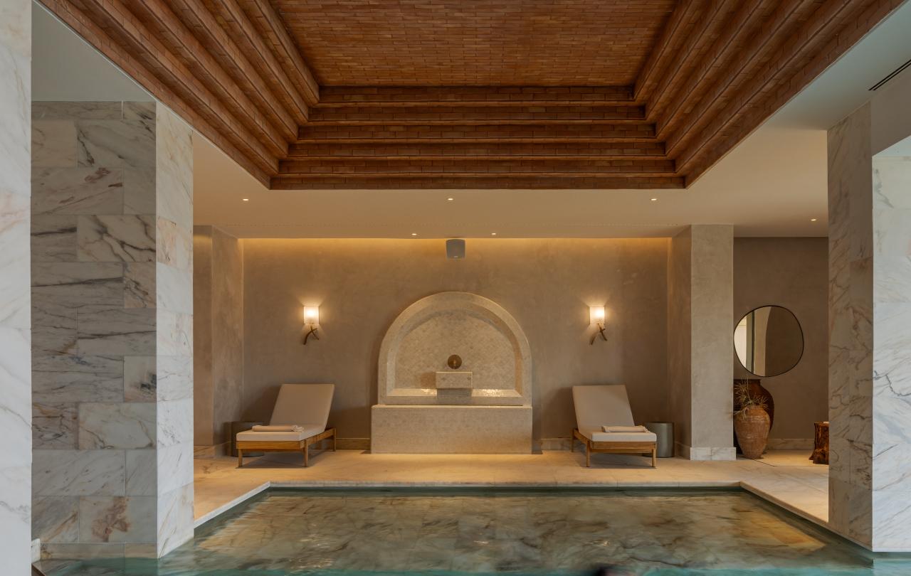 Uma pessoa nada numa piscina coberta, sob um teto de madeira em degraus, com duas espreguiçadeiras e uma fonte visíveis no fundo da sala.