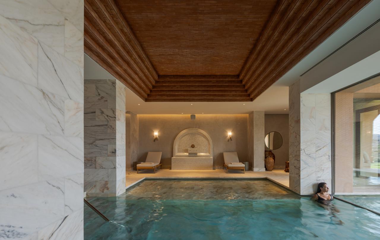 Piscina interior com água turquesa sob um teto de madeira em degraus, com colunas de mármore e duas espreguiçadeiras ao fundo.