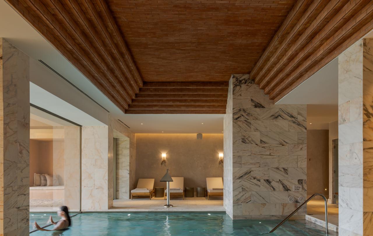 Uma piscina interior com águas turquesa cristalinas e uma pessoa a relaxar nela, situada num spa de luxo que apresenta um teto de madeira em camadas característico, colunas de mármore e espreguiçadeiras ao fundo.