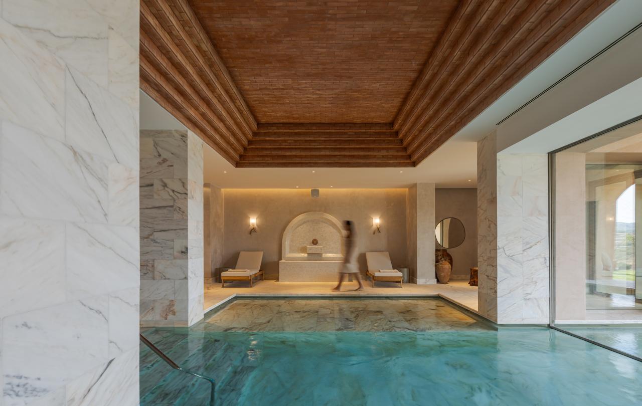 Uma grande piscina interior com águas cristalinas de cor turquesa e um pavimento revestido a mármore, sob um teto de tábuas de madeira, com duas espreguiçadeiras ao fundo.