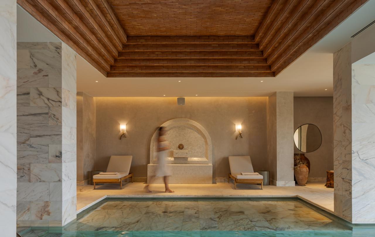 Uma luxuosa piscina interior e sala de spa com um teto de madeira em camadas, pilares de mármore, espreguiçadeiras e uma pessoa a caminhar ao fundo.