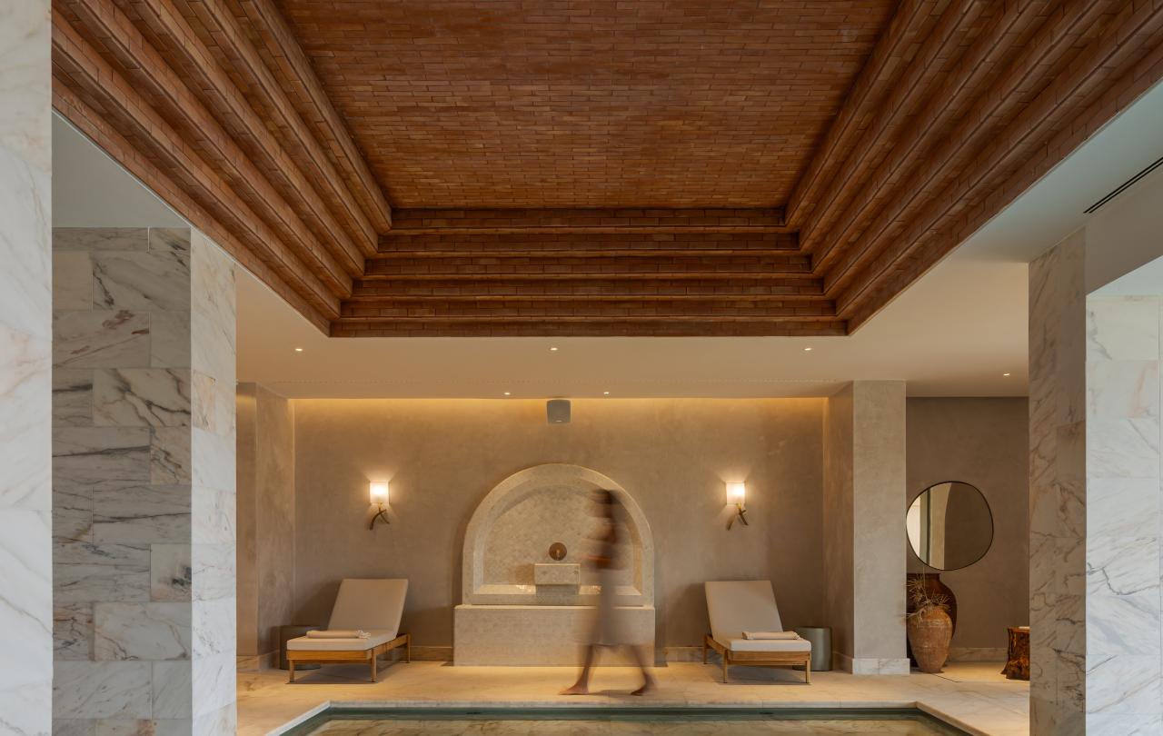 Spa interior com uma piscina de águas turquesa e espreguiçadeiras sob um teto de madeira em camadas, com uma pessoa em movimento, com a imagem desfocada, ao fundo.