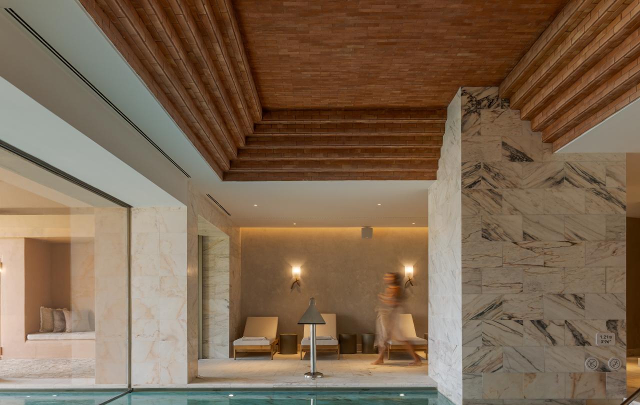 Uma piscina interior de luxo apresenta um teto em degraus de tijolo vermelho, pilares de mármore e uma área de estar com assentos ao fundo.