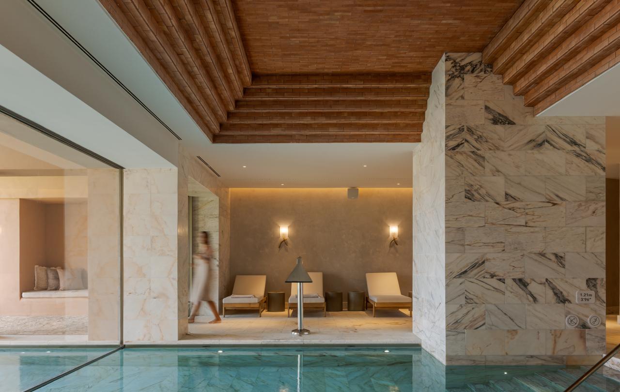 Piscina interior com água azul-clara e revestimento em mármore, situada numa sala com um teto decorativo em tijolo e três espreguiçadeiras ao fundo.