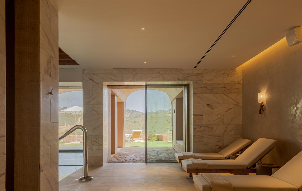Sala de relaxamento do spa com iluminação acolhedora, com filas de espreguiçadeiras viradas para uma grande porta de correr de vidro que se abre para uma paisagem exterior ensolarada.