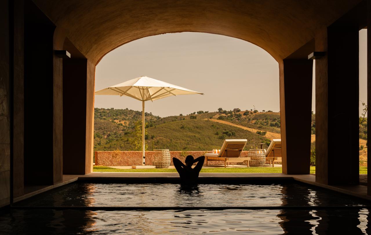 Uma pessoa relaxa numa piscina interior-exterior com vista para colinas verdes ondulantes, espreguiçadeiras e um guarda-sol junto a uma abertura em arco.