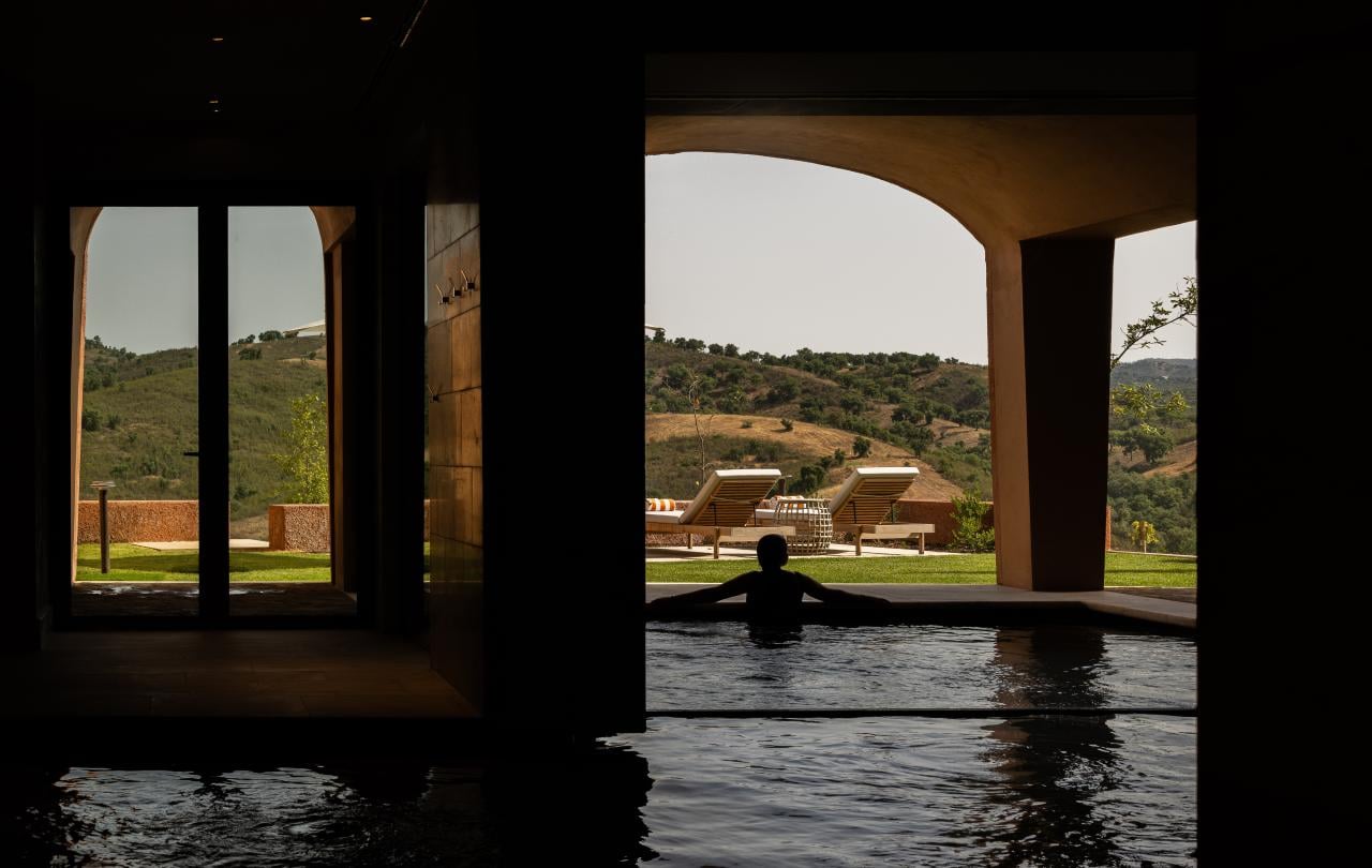 Silhueta de uma pessoa numa piscina coberta, a olhar por uma abertura em arco na direção de colinas verdes e ensolaradas.