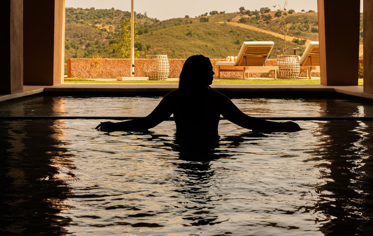 Silhueta de uma pessoa a flutuar numa piscina, emoldurada por um grande arco com vista para as colinas.