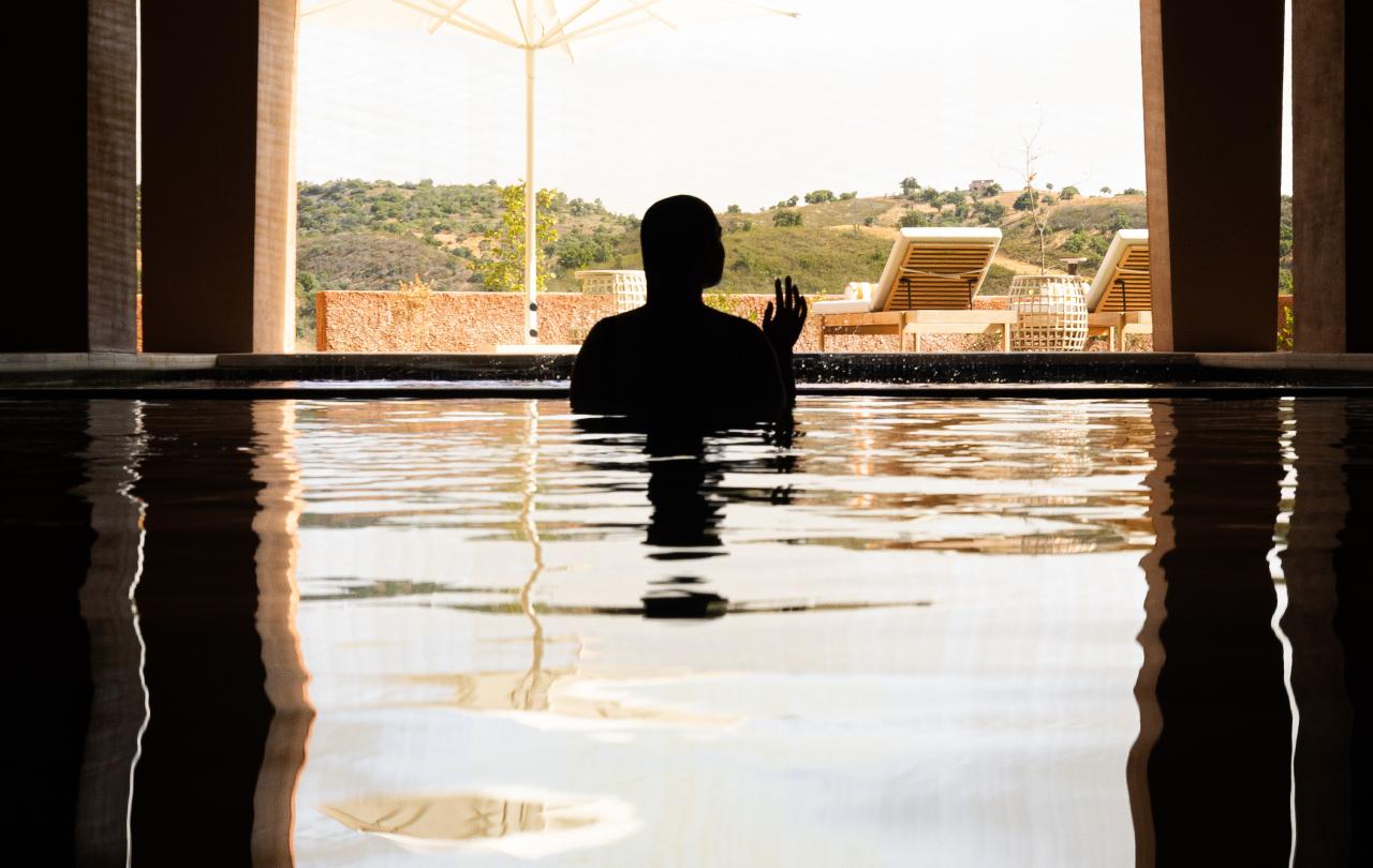 Silhueta de uma pessoa a relaxar numa piscina interior, emoldurada por uma grande janela em arco com vista para um terraço exterior ensolarado, com espreguiçadeiras e colinas ondulantes.