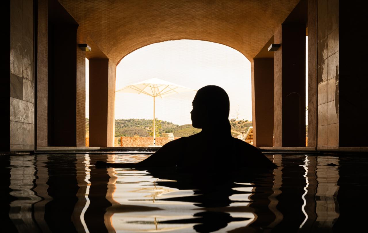 Silhueta de uma pessoa numa piscina a olhar através de uma passagem em arco banhada pela luz do sol.