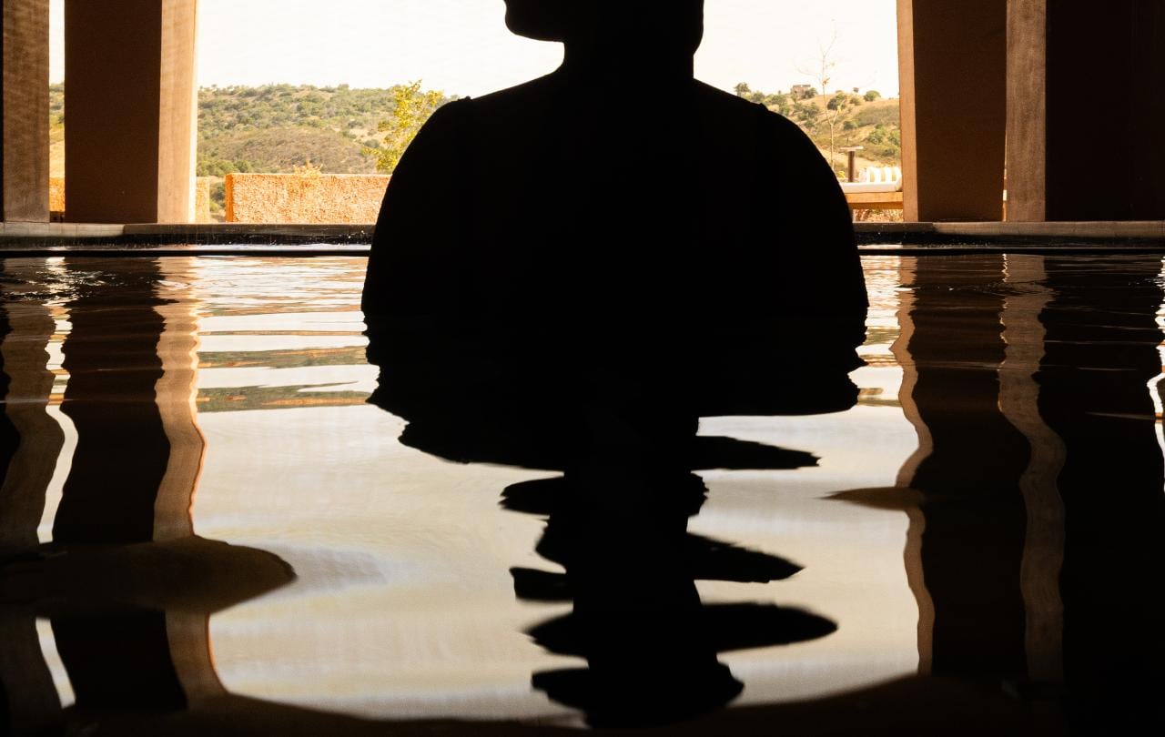 Silhueta de uma pessoa de pé numa piscina, sob um arco banhado pelo sol, com o seu reflexo a cintilar na superfície da água.