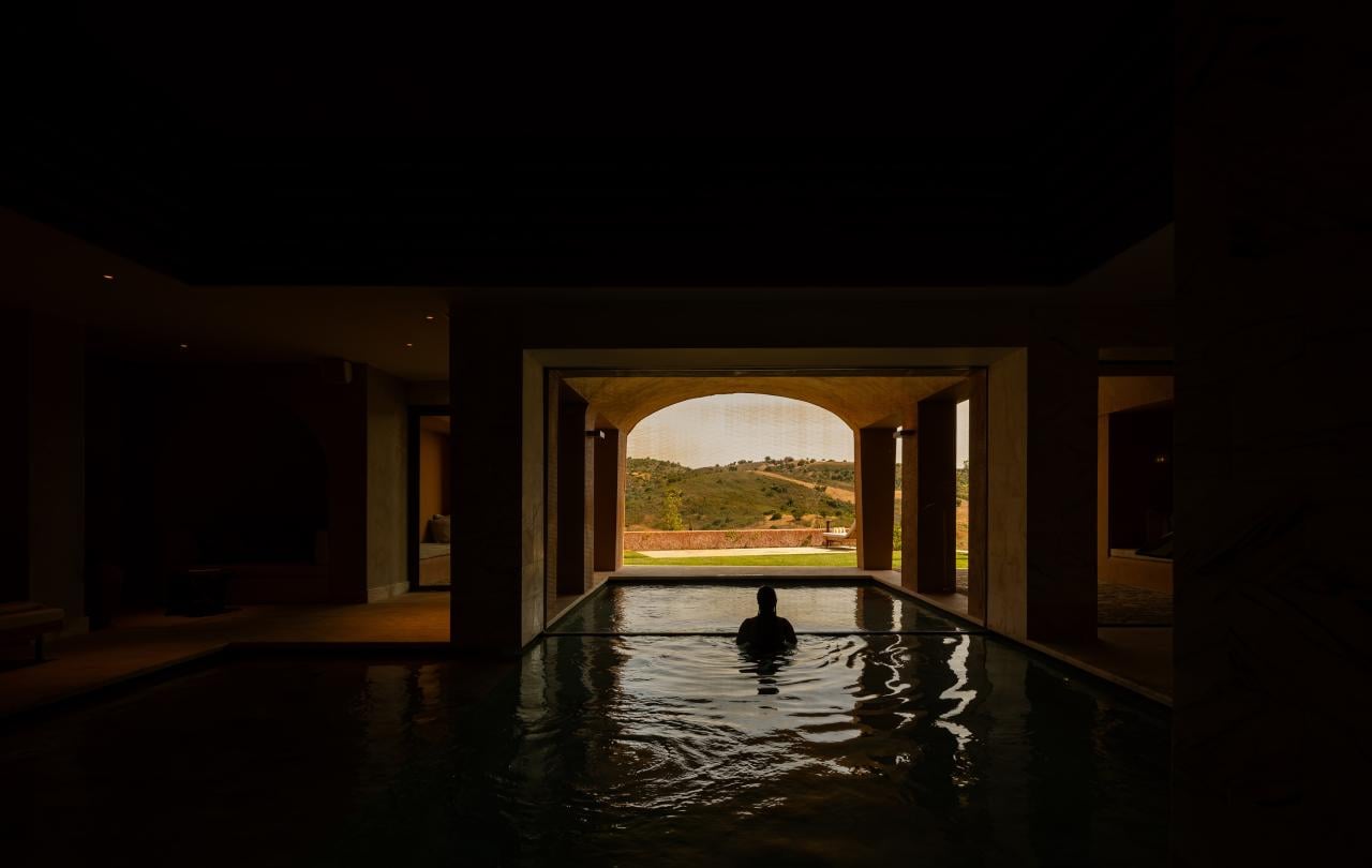 Vista ampla de uma piscina interior pouco iluminada que conduz a um arco exterior bem iluminado.