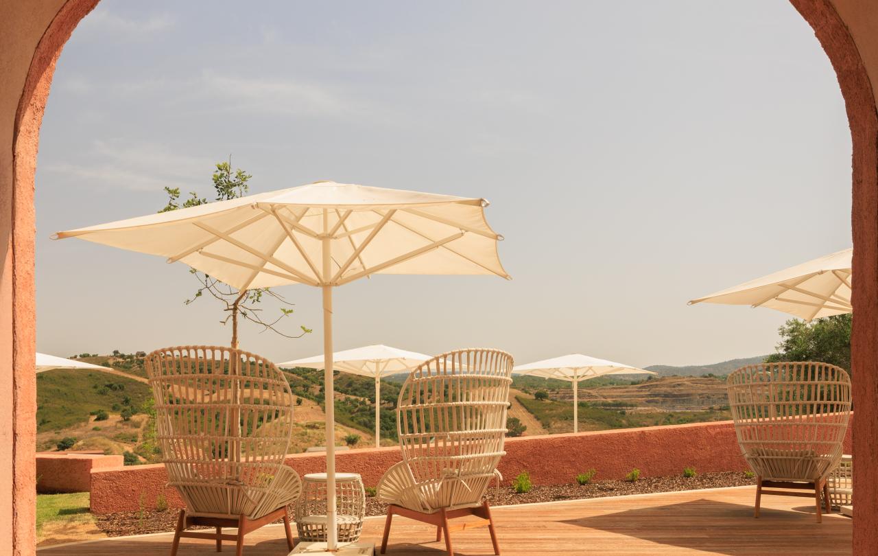 Terraço exterior moderno, com poltronas de vime e guarda-sóis, com vista para colinas ondulantes.