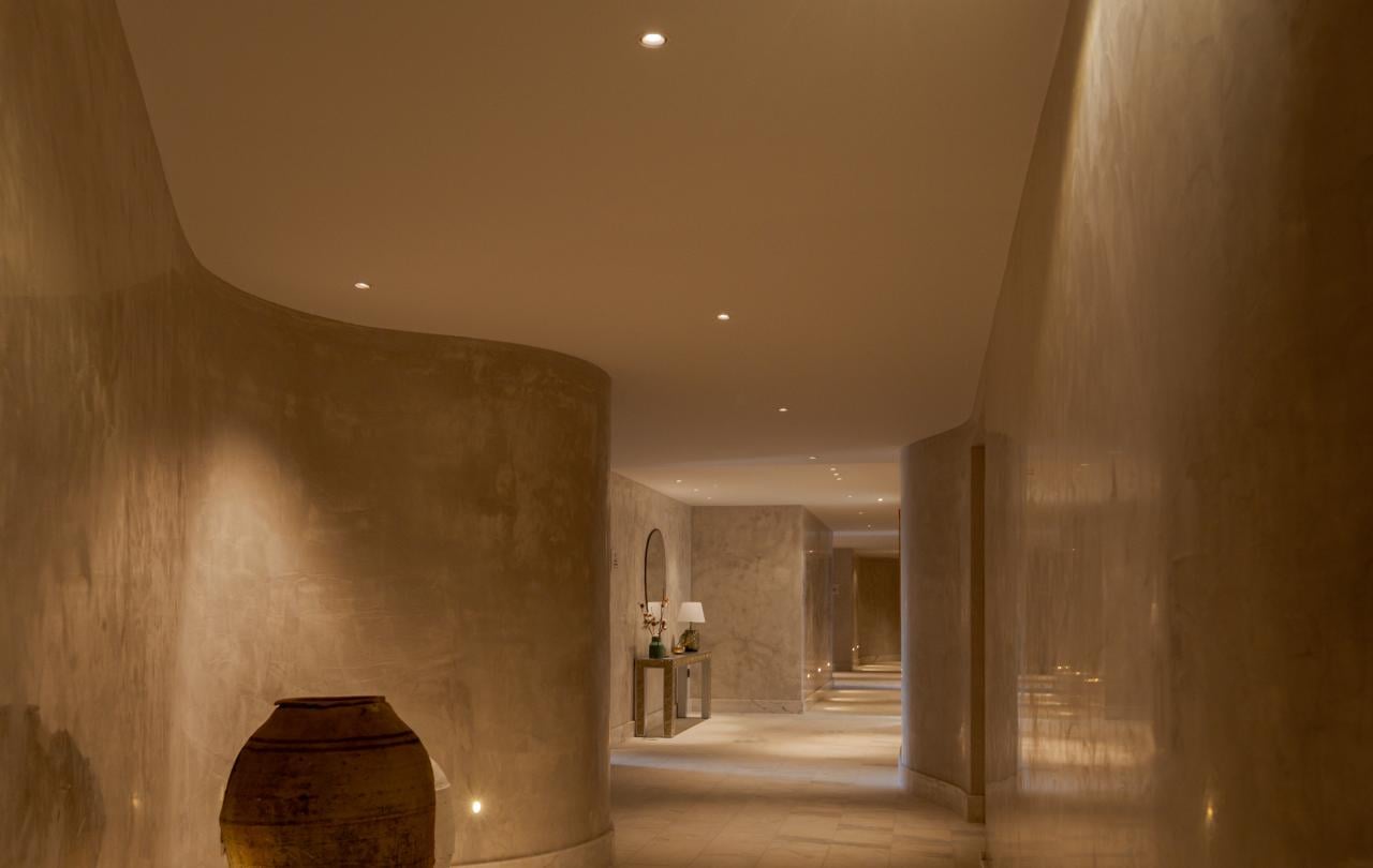 Corredor minimalista de spa com paredes quentes e texturadas, pavimentos polidos e uma grande urna decorativa.