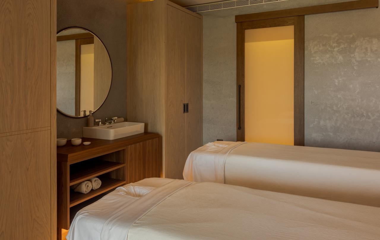 Sala de tratamentos de spa com duas camas de massagem, uma penteadeira em madeira e um espelho circular.