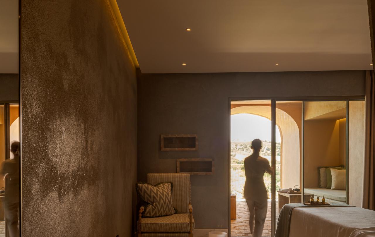 Uma terapeuta de spa em pé junto a portas de vidro abertas numa sala de tratamentos relaxante com vista para a varanda.
