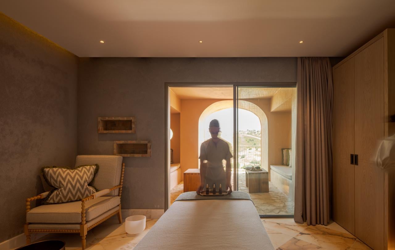 Uma terapeuta de spa a preparar óleos numa marquesa de massagem, numa sala de tratamentos tranquila com varanda.