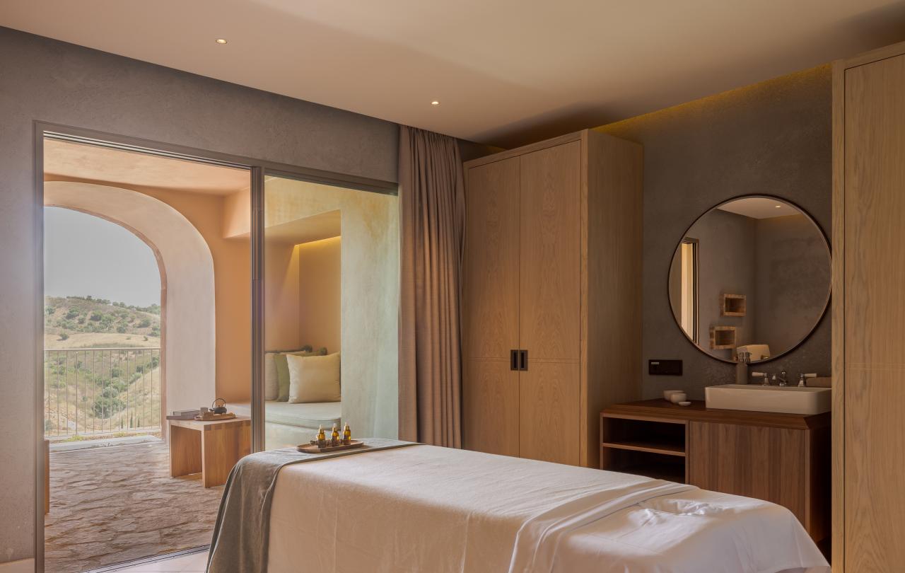 Sala de tratamentos de spa luminosa, com uma marquesa de massagem preparada e uma porta de vidro de correr que dá acesso a uma varanda.